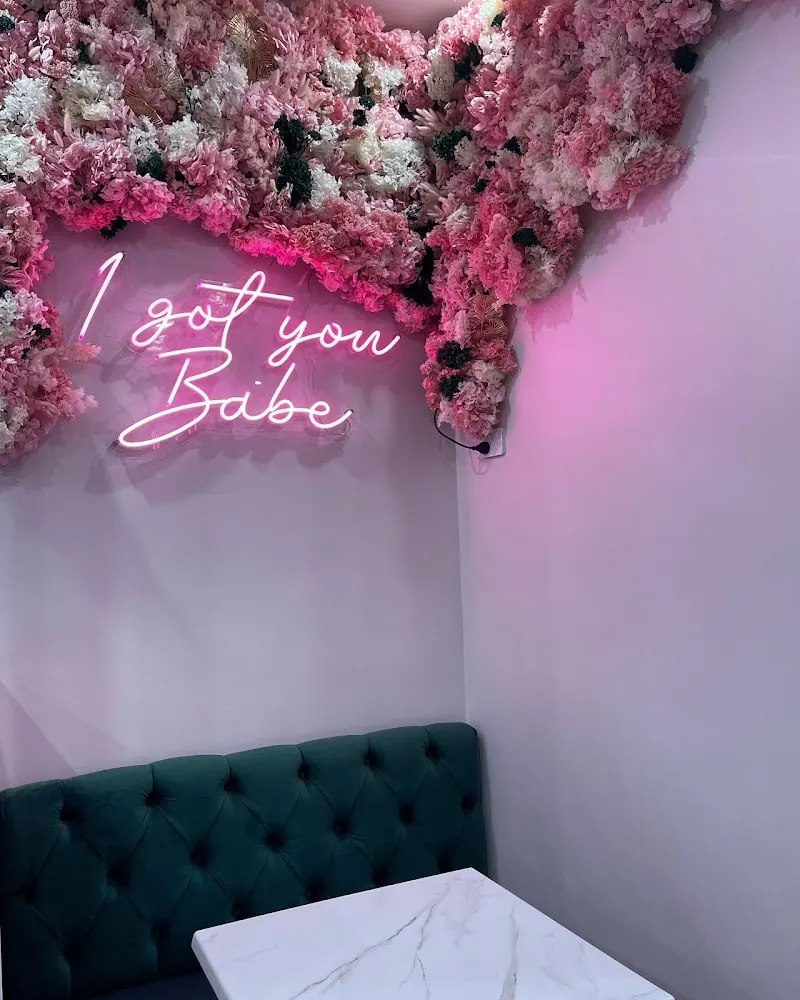 A teal velvet booth and marble table under the flower wall and a pink 'I got you babe' neon sign